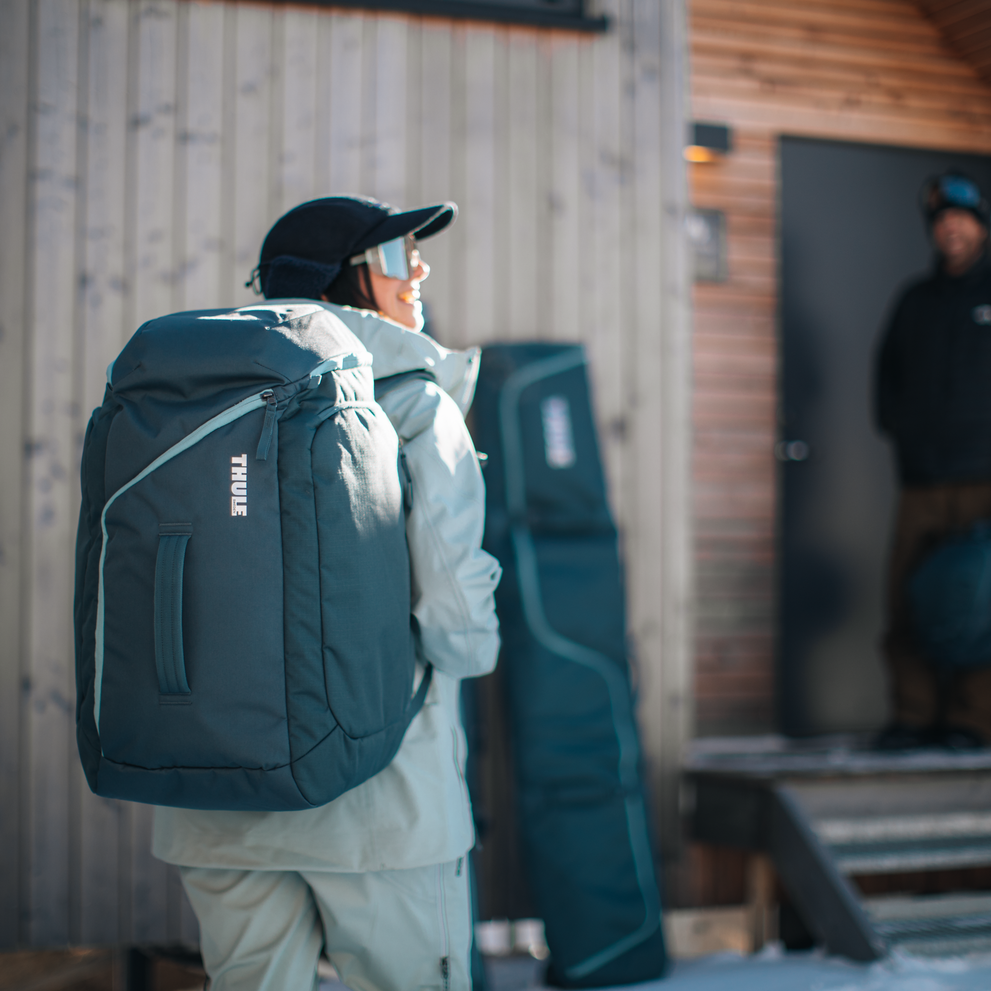 A man is standing outside a house carrying a Thule RoundTrip Ski and Snowboard Boot Backpack.