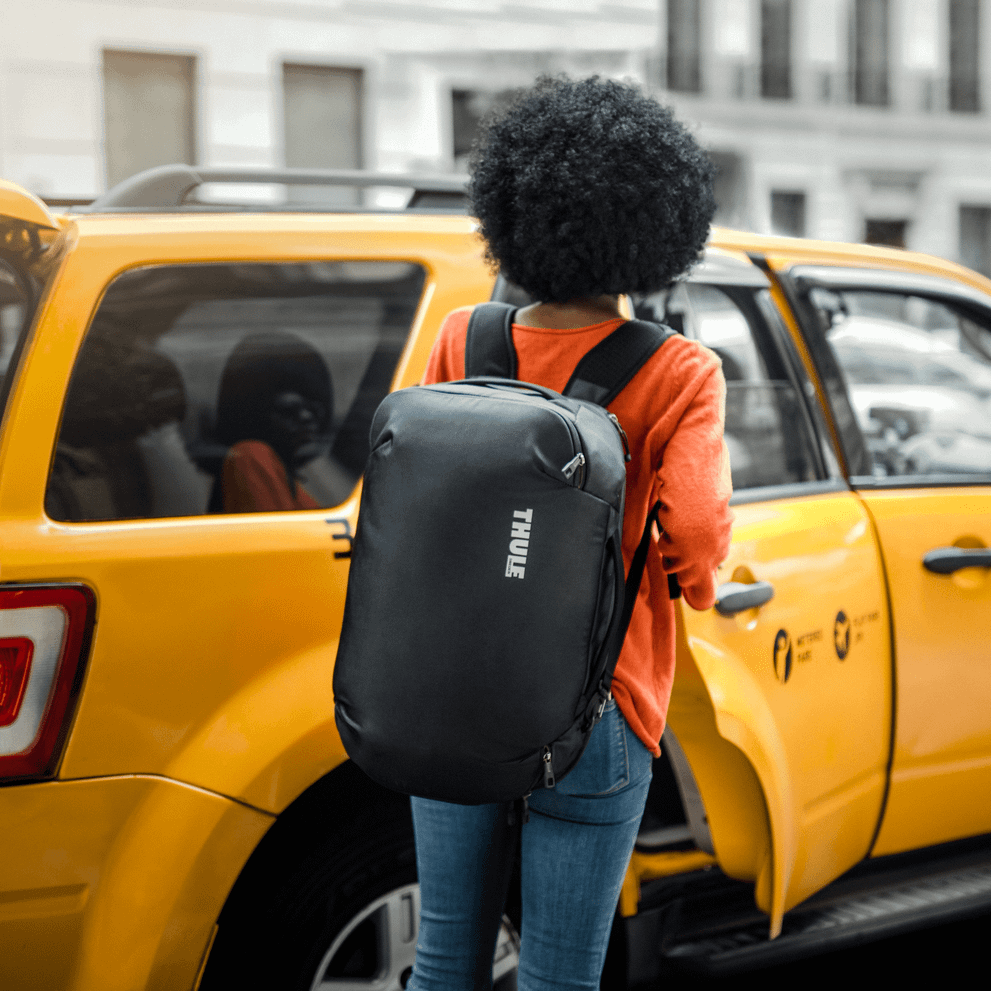 A woman enters a yellow taxi carrying a Thule Subterra Convertible Carry-On backpack.