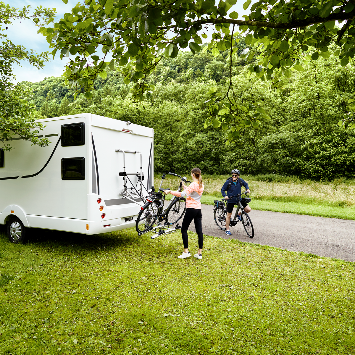 A motorhome parked in the grass and people unload bikes from a Thule Lift V16  rv bike rack.