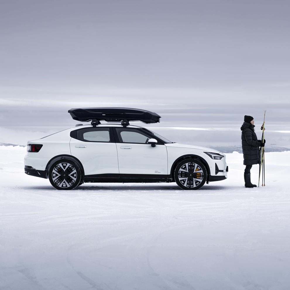 A woman is standing in front of a car with a Thule Motion 3, holding a pair of skis, in a snowy landscape,