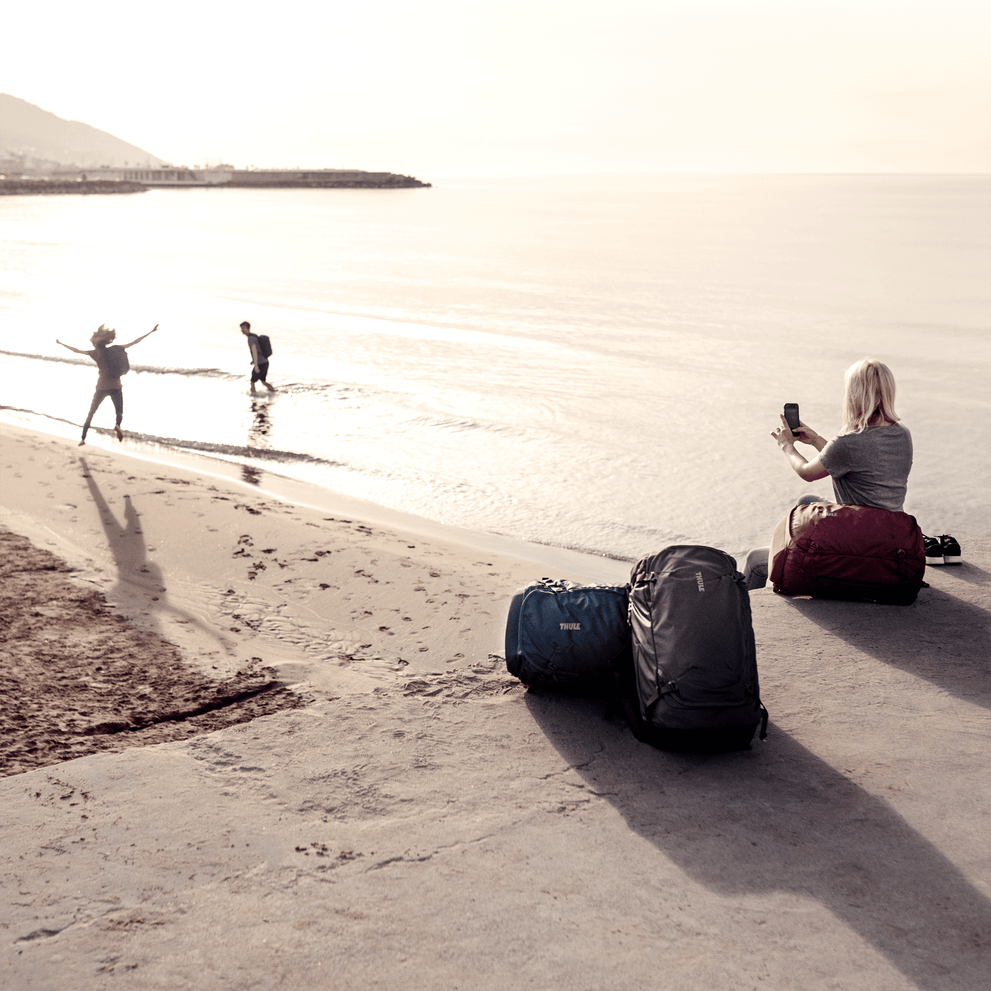 A woman with a a Thule Landmark 60L travel backpack sits next to the beach taking a picture of her friends.