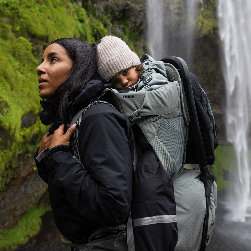 Next to a waterfall, a woman goes for a hike with her toddler in a child carrier backpack.