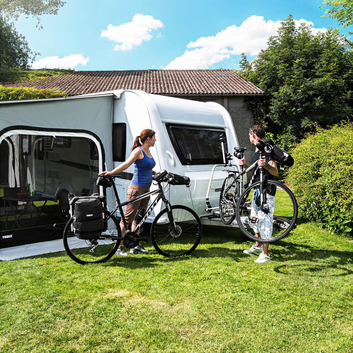 Two cyclists stand behind a caravan that has bikes on the Thule Caravan Superb Standard caravan bike rack.