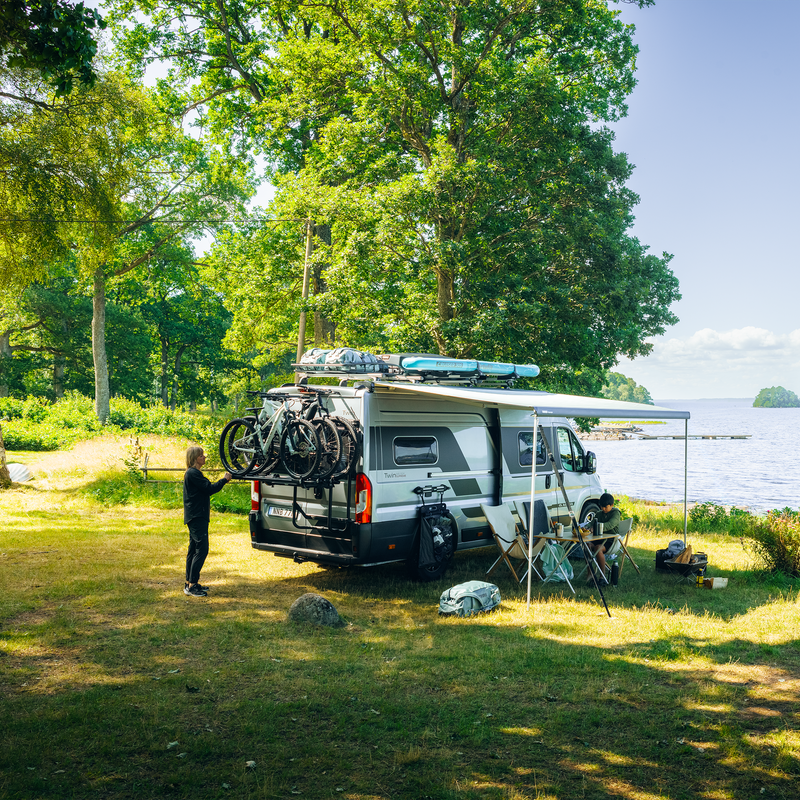 A camper van parked at a scenic lakeside campsite, featuring the Thule VeloTrack 3rd Rail Kit carrying three bikes. A person prepares gear while the awning provides shade, creating a comfortable outdoor experience.