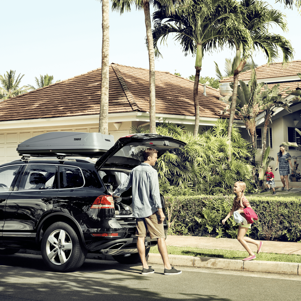 A man is standing by a car with the trunk open and a Thule Touring roof box mounted, while a girl is approaching the car