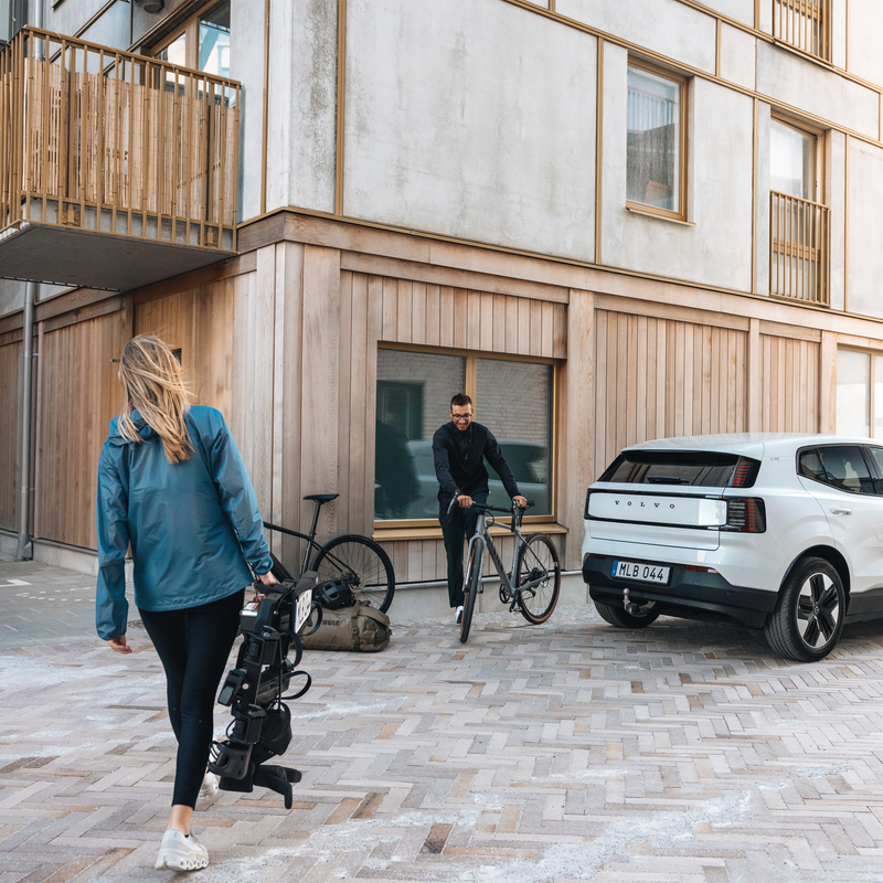 A woman carrying a folded Thule OutPace bike rack while a man approaches a parked car with two bikes.