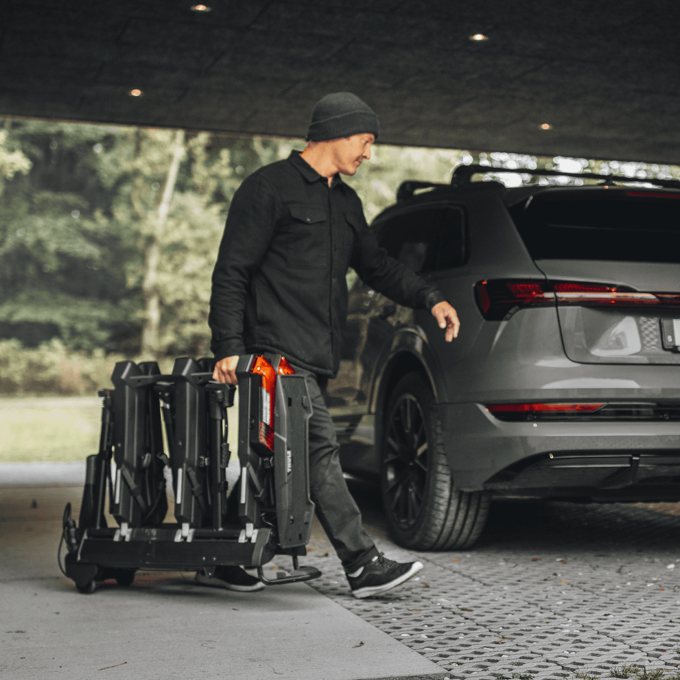 A man walks through a garage towards his car with the folded Thule Epos hitch bike rack rolling on wheels.