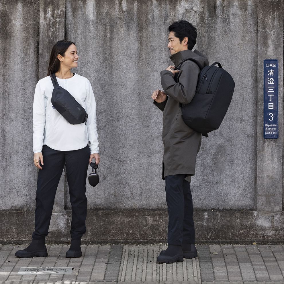 A man and woman stand next to a concrete wall, talking and carrying a Thule Subterra backpack and crossbody.