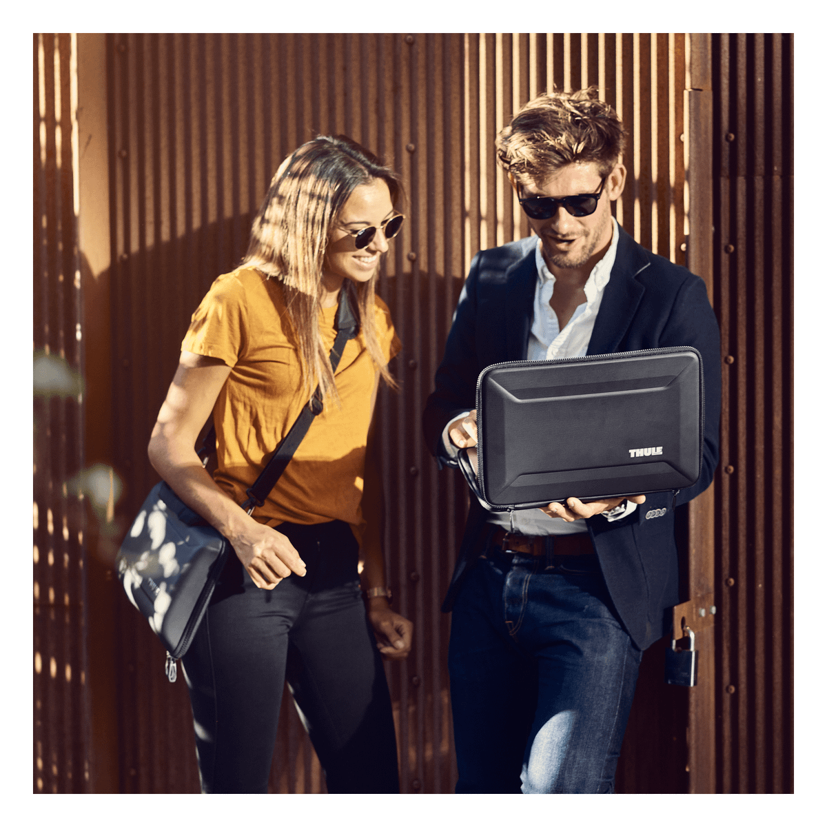 Leaning against a brown wall in the sunshine a man shows a woman his laptop inside a black Thule Gauntlet 4 MacBook Pro Attaché.