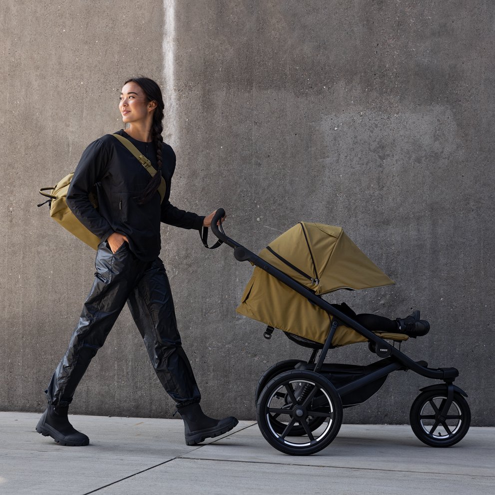 A woman walks down the street with a green backpack and a green Thule Urban Glide 3 all-terrain stroller.
