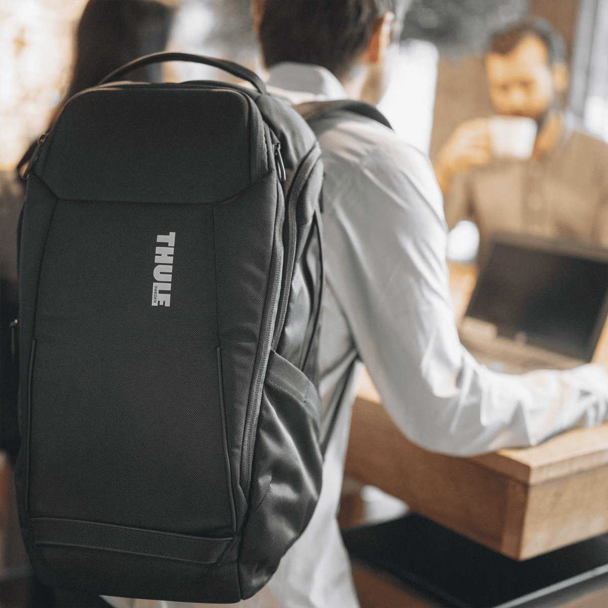 A person sits at a cafe table with a laptop and a black Thule Accent backpack.
