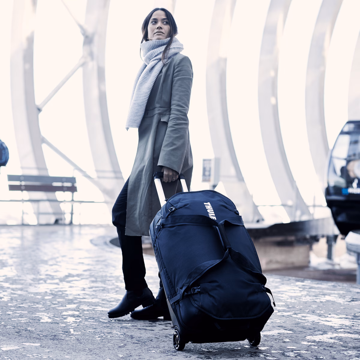 In a cold bus station, a woman waits with a scarf and her Thule Subterra Wheeled Duffel suitcases.
