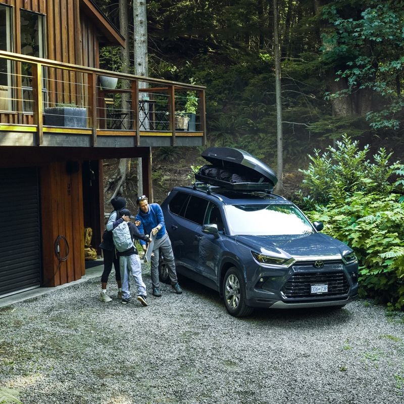 A family is standing near a car with an open Thule Force 3 rooftop cargo box on its roof, parked outside a modern wooden house surrounded by lush forest.
