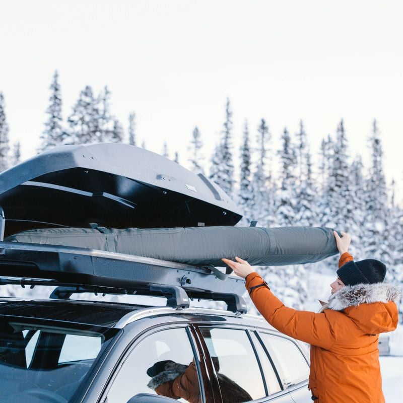 A woman in an orange ski jacket in a snowy setting loads a Thule RoundTrip ski bag into the roof box of a car.