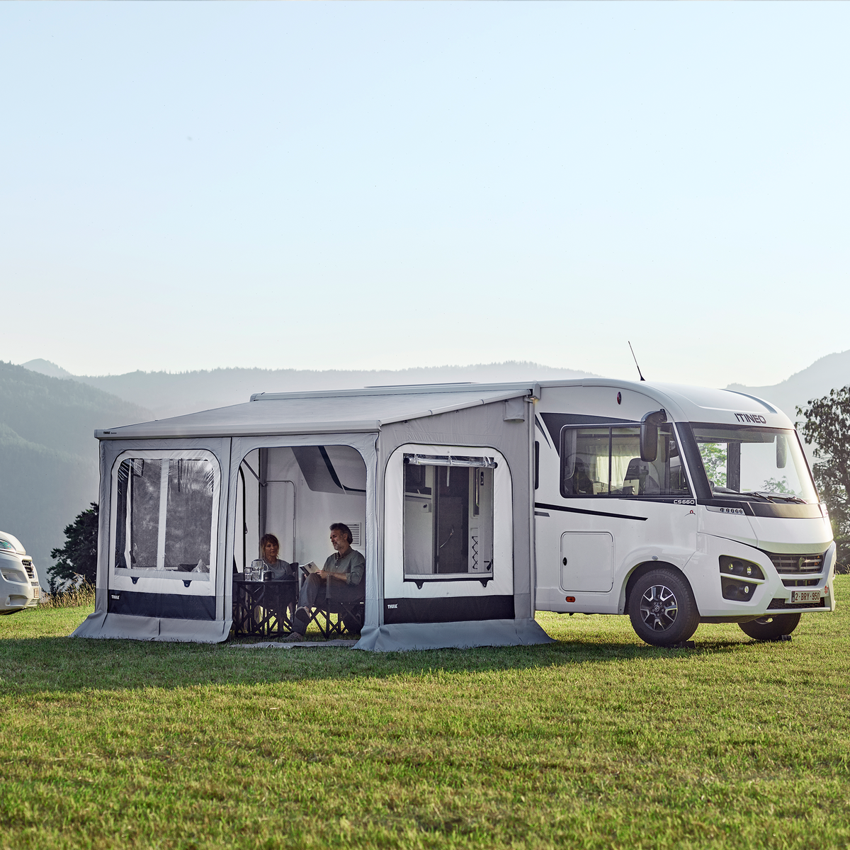 A motorhome with a Thule Panorama rv tent is parked in a field next to a van with a pop-up roof.