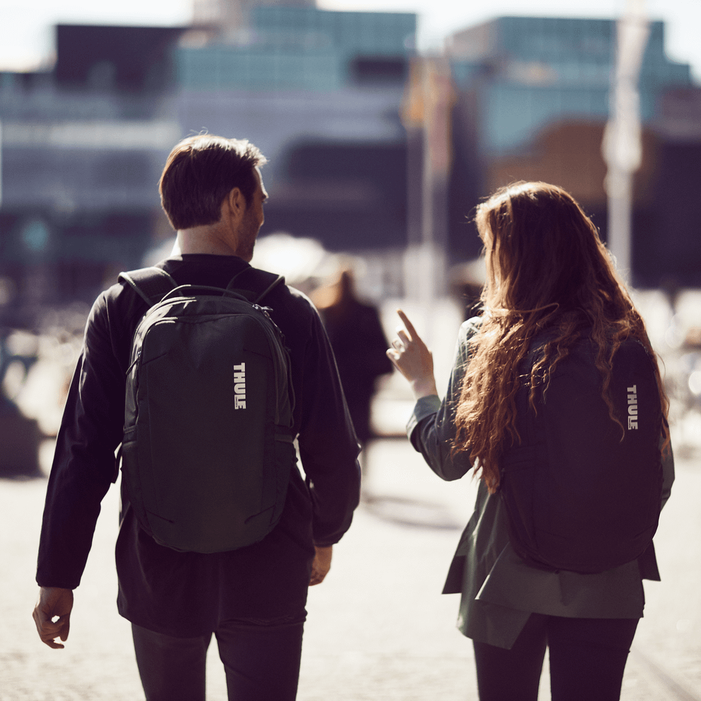 Two people walk on a city square with glass buildings in the background, holding Thule Subterra backpacks.