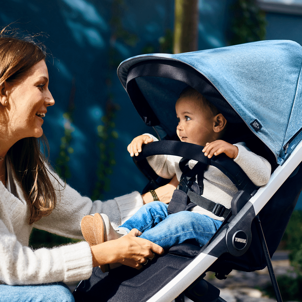 A woman smiles at her baby in a stroller with a blue Thule Spring Canopy.