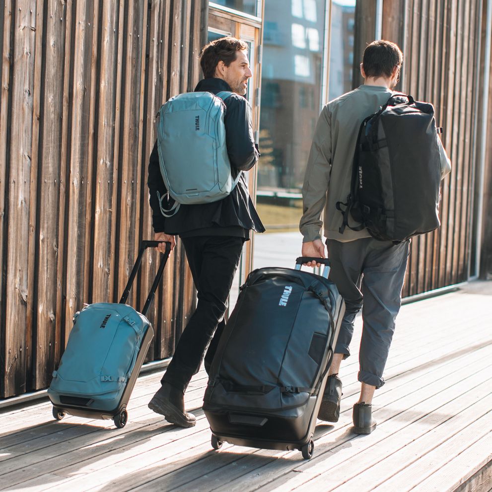 Two men are walking down a sunny path with black and blue Thule Chasm duffels, backpacks and suitcases.
