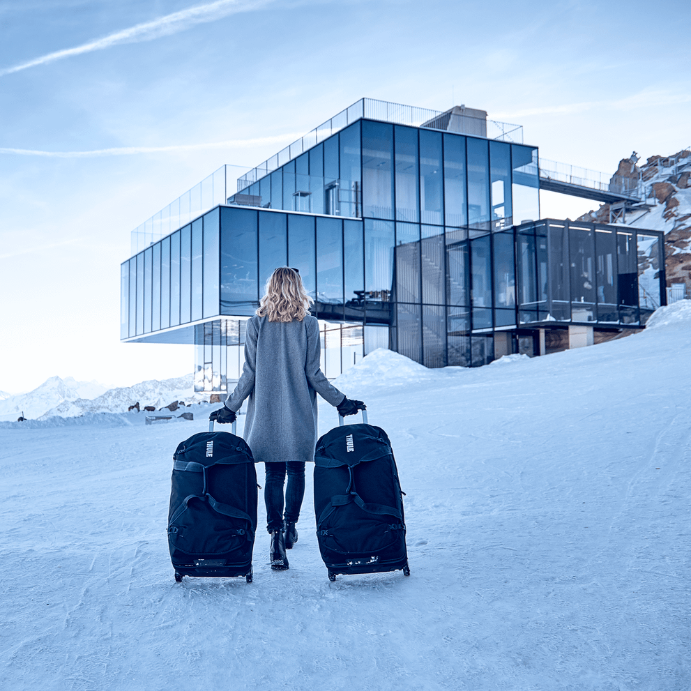 In the snow, a woman walks towards a glass building with two Thule Subterra Wheeled Duffel suitcases.