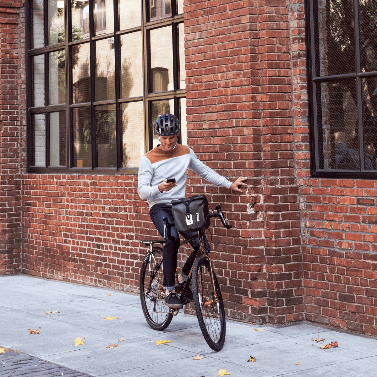 A man sits on his bike with a Thule Shield Handelbar bag, leaning against a brick wall, looking at his phone.