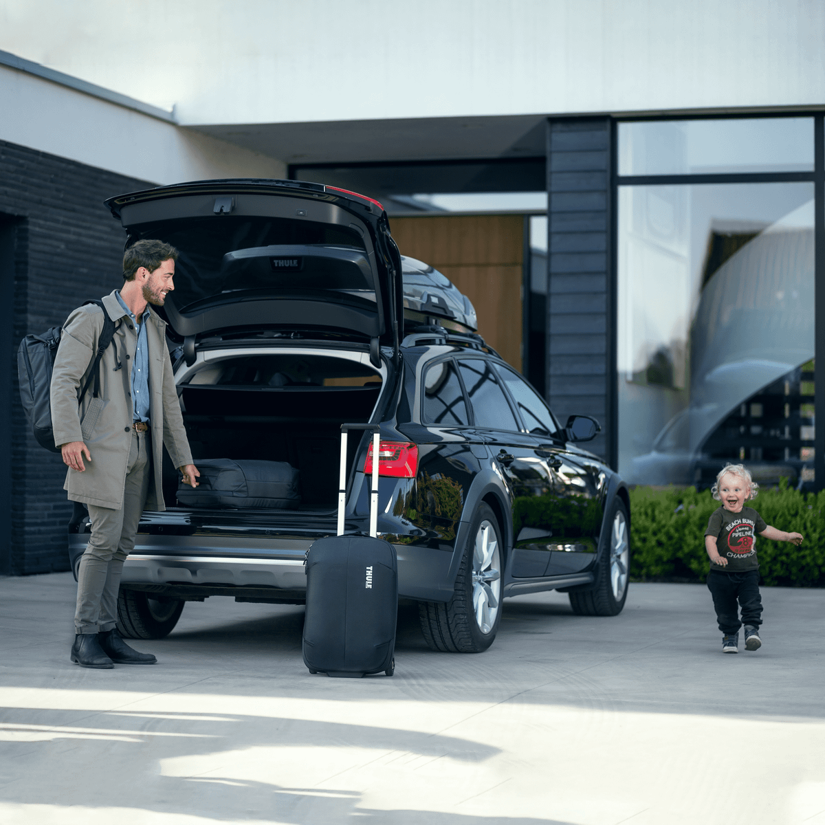 A man opens the trunk of his car, and a kid runs around as he takes out his Thule Subterra Carry-On suitcase.