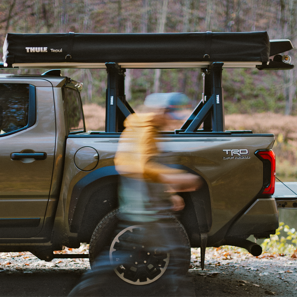 A man walks beside a vehicle with Thule Xscape truck rack that carries the rooftop tent Thule Foothill and the fishing rod rack Thule RodVault 2 in an outdoors environment.
