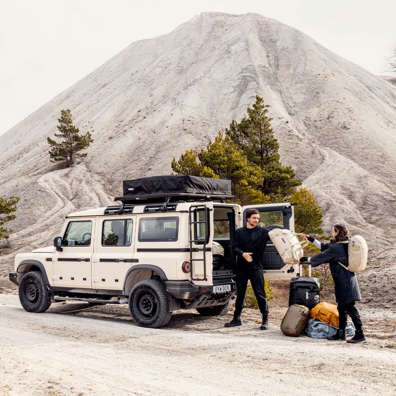 A man with short dark hair and a woman with long brown hair are unloading bags of various colors and sizes from the open trunk of a Jeep. They are standing in a rugged outdoor environment uneven ground and sparse vegetation.
