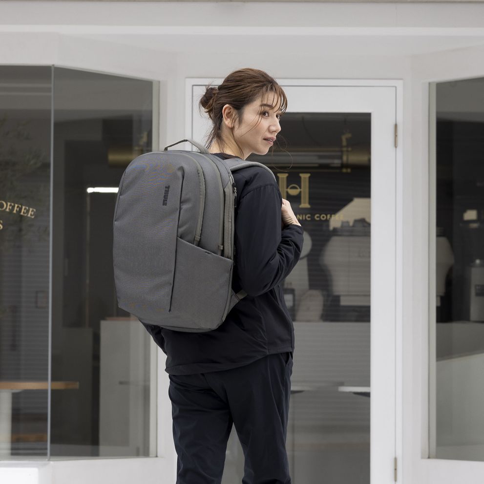 A woman stands outside of a white building with glass doors, holding a gray Thule Subterra backpack.