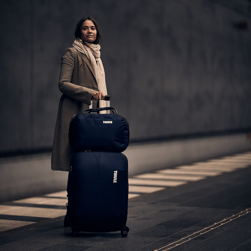 A woman looks outside a window at the sunshine with a suitcase and Thule Subterra Boarding Bag.