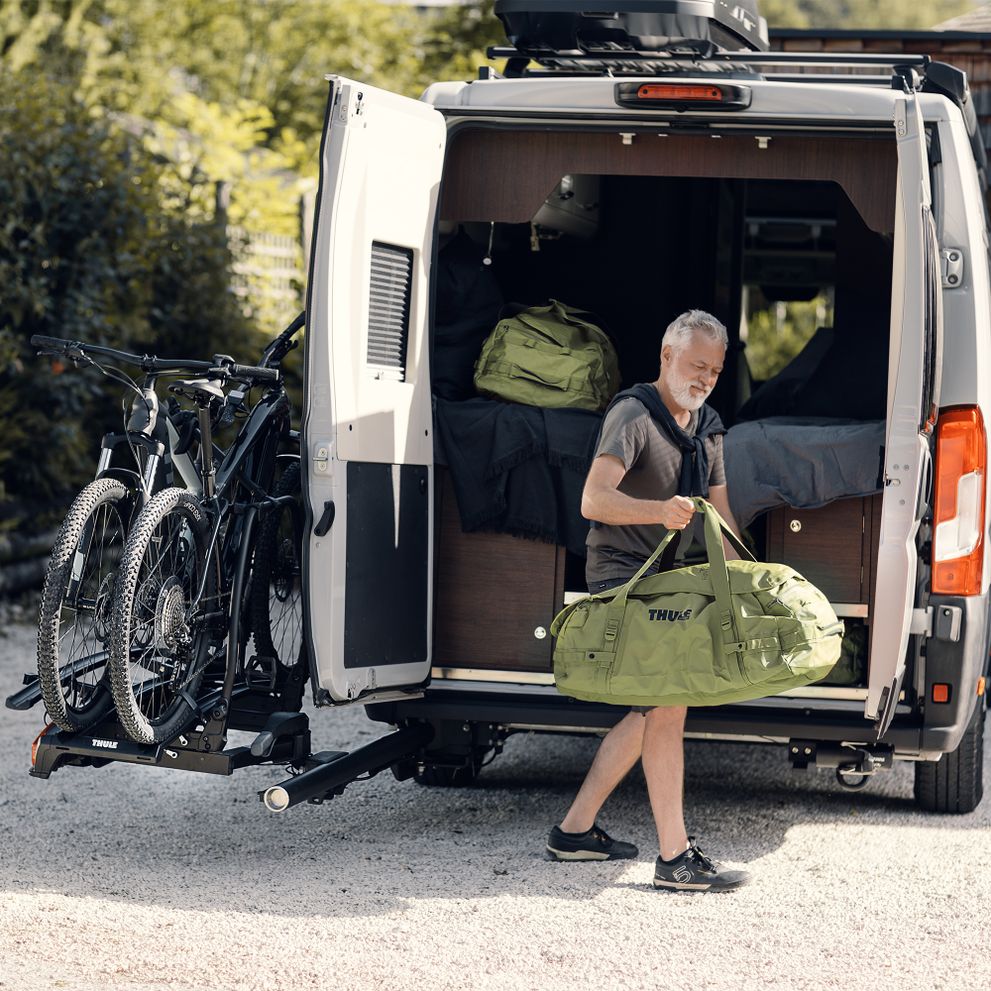 A man loads gear from the back of his white van that has the trunk door open and a swingable Thule VeloSwing towbar with bike rack.