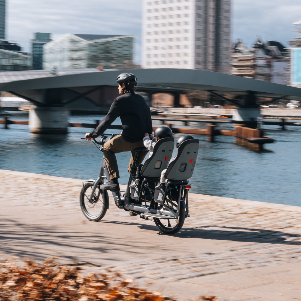 Person, die mit einem Fahrrad fährt, das mit einem Thule Yepp Nexxt 2 Kindersitz ausgestattet ist, entlang eines Ufers mit einer Stadtbrücke und Gebäuden im Hintergrund.