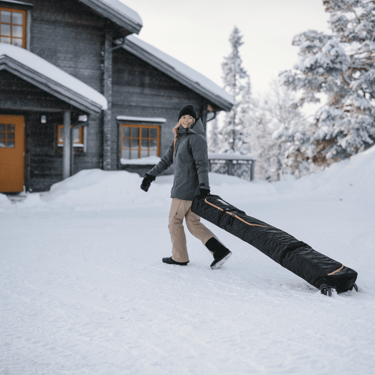 A woman stands in the snow outside a cottage wheeling her Thule RoundTrip travel snowboard bag.