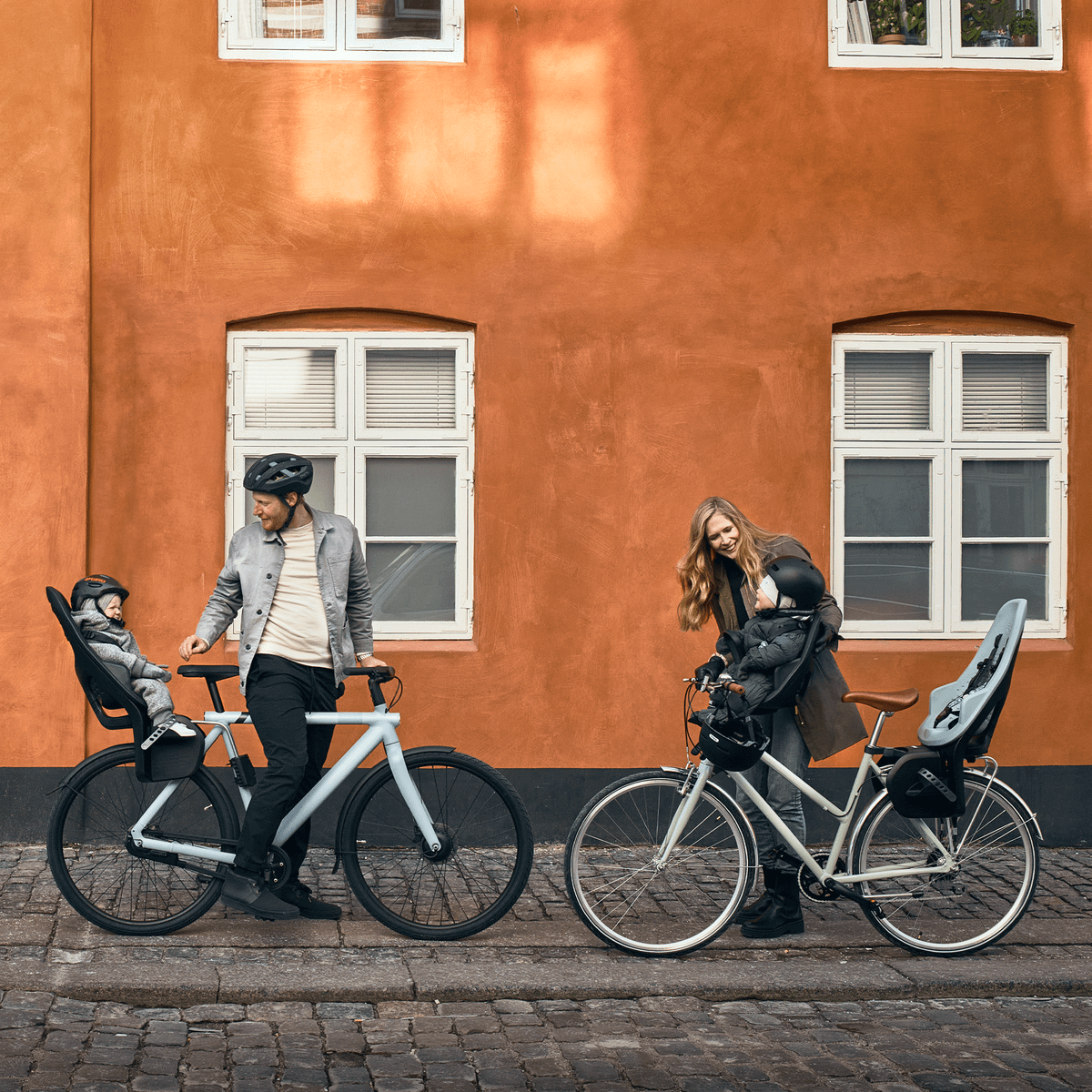 Next to a orange wall, two parents stand with bikes and kids in Thule Yepp 2 Mini and Maxi child bike seats.