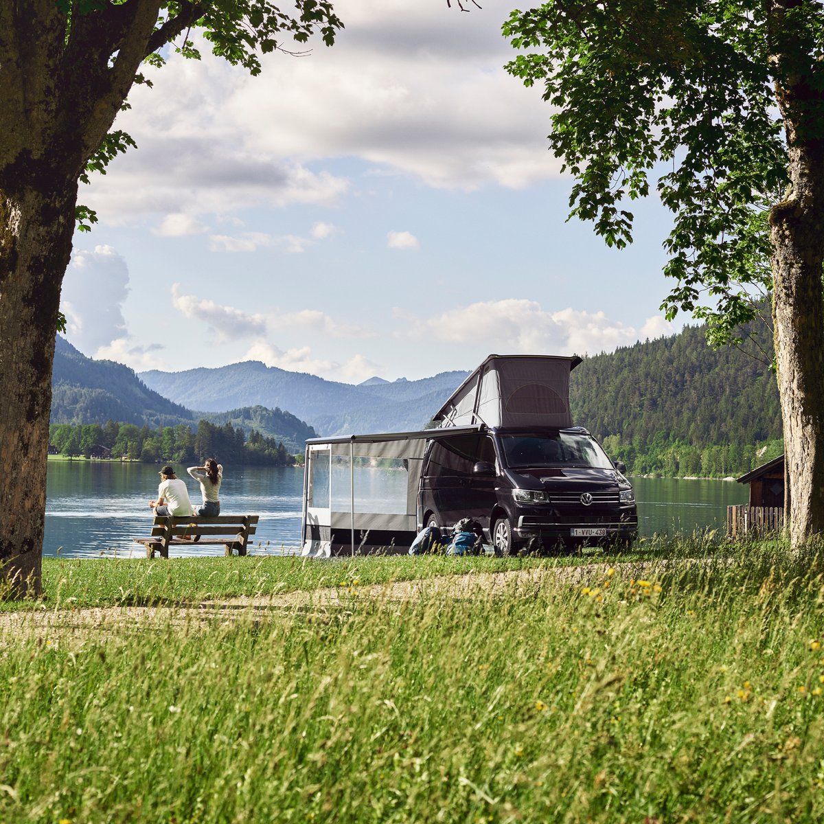 A black van is parked near a lake with a pop-up roof and a black Thule Hideaway awning