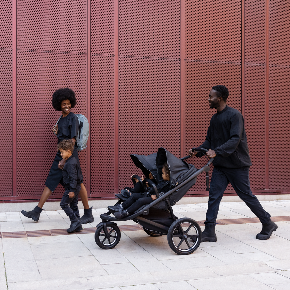 A family walks down a street with the father pushing two kids in the black Thule Urban Glide double stroller.