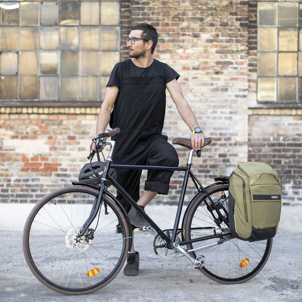 A man stands with his bike by a brick wall with a green Thule Paramount bike pannier.