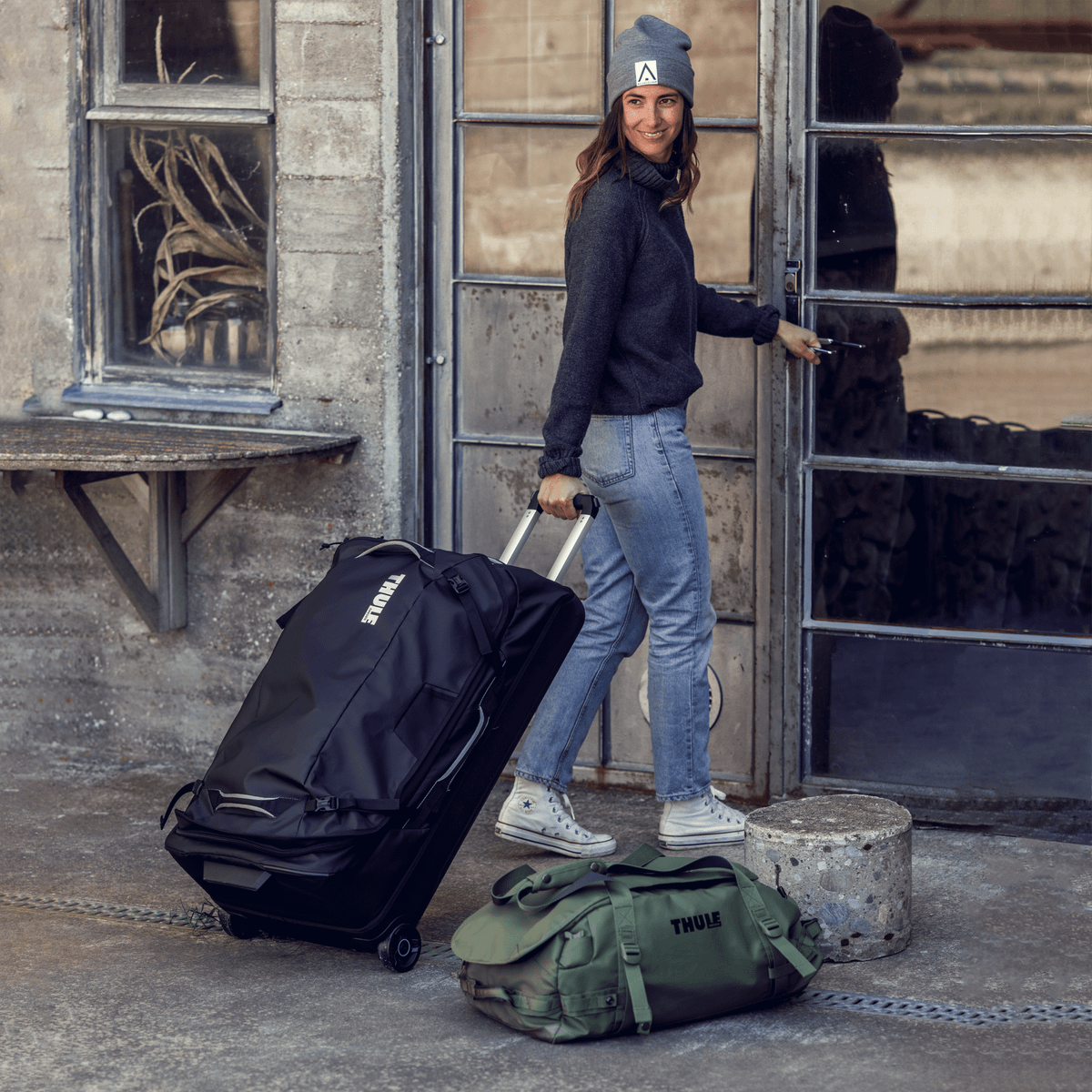 A woman walks into a concrete building with a green duffel and  black Thule Chasm suitcase.