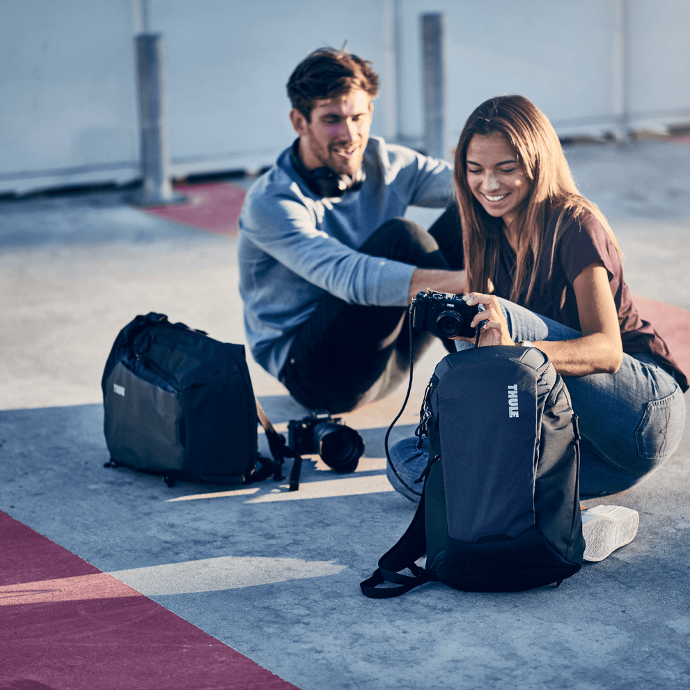 Two people sit on the ground and put their cameras into black Thule EnRoute Medium DSLR camera backpacks.