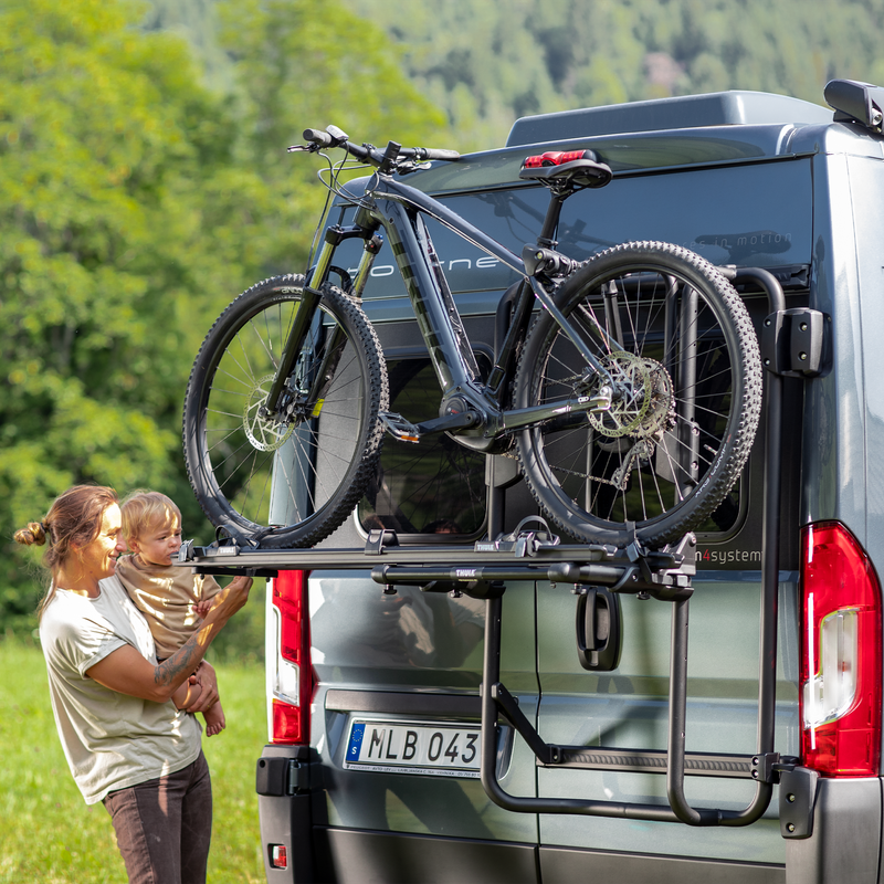 A woman effortlessly secures a mountain bike onto the Thule VeloTrack bike rack mounted on the rear of a van. The sturdy and spacious design allows for easy loading of large bikes.
