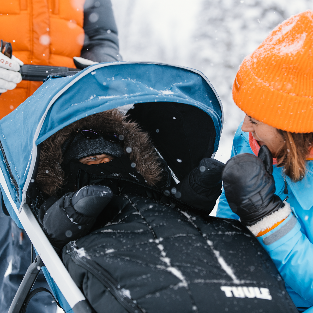 A baby sits in a jogging stroller in the snow with a Thule Footmuff Sport.