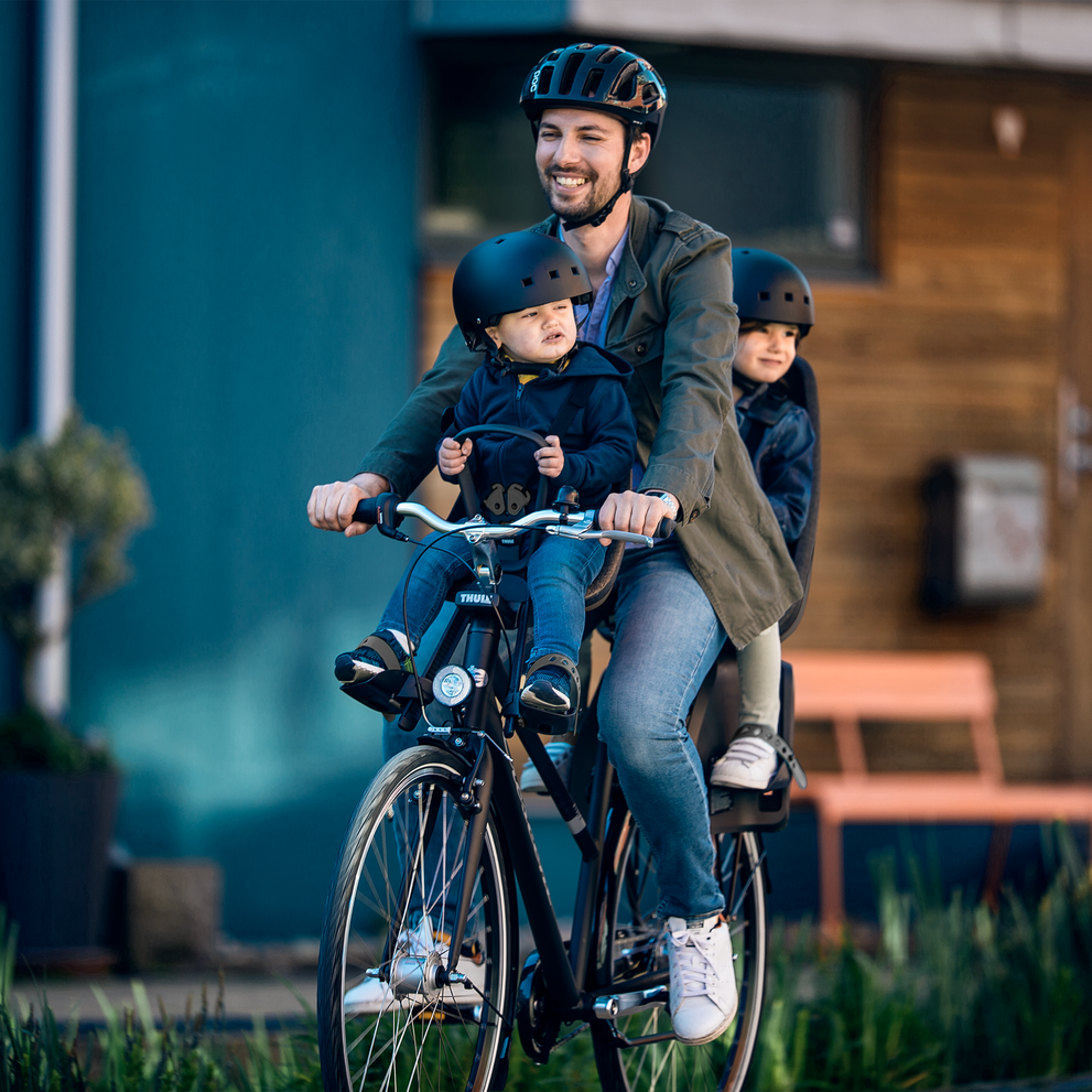 A man bikes down a bike path with his kids in kid's bike seats, including the Thule Yepp Nexxt Mini bike seat.