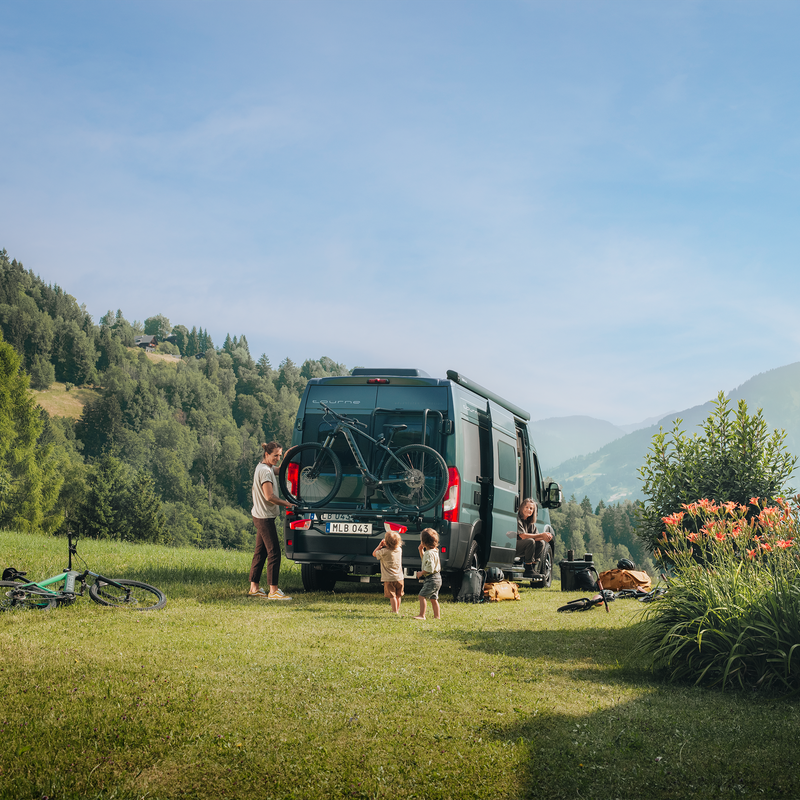 A camper van set up in a scenic outdoor location, featuring the Thule VeloTrack with the Thule Lightboard Van. The bikes are mounted at a lower position, ensuring taillights remain visible while making loading easier.