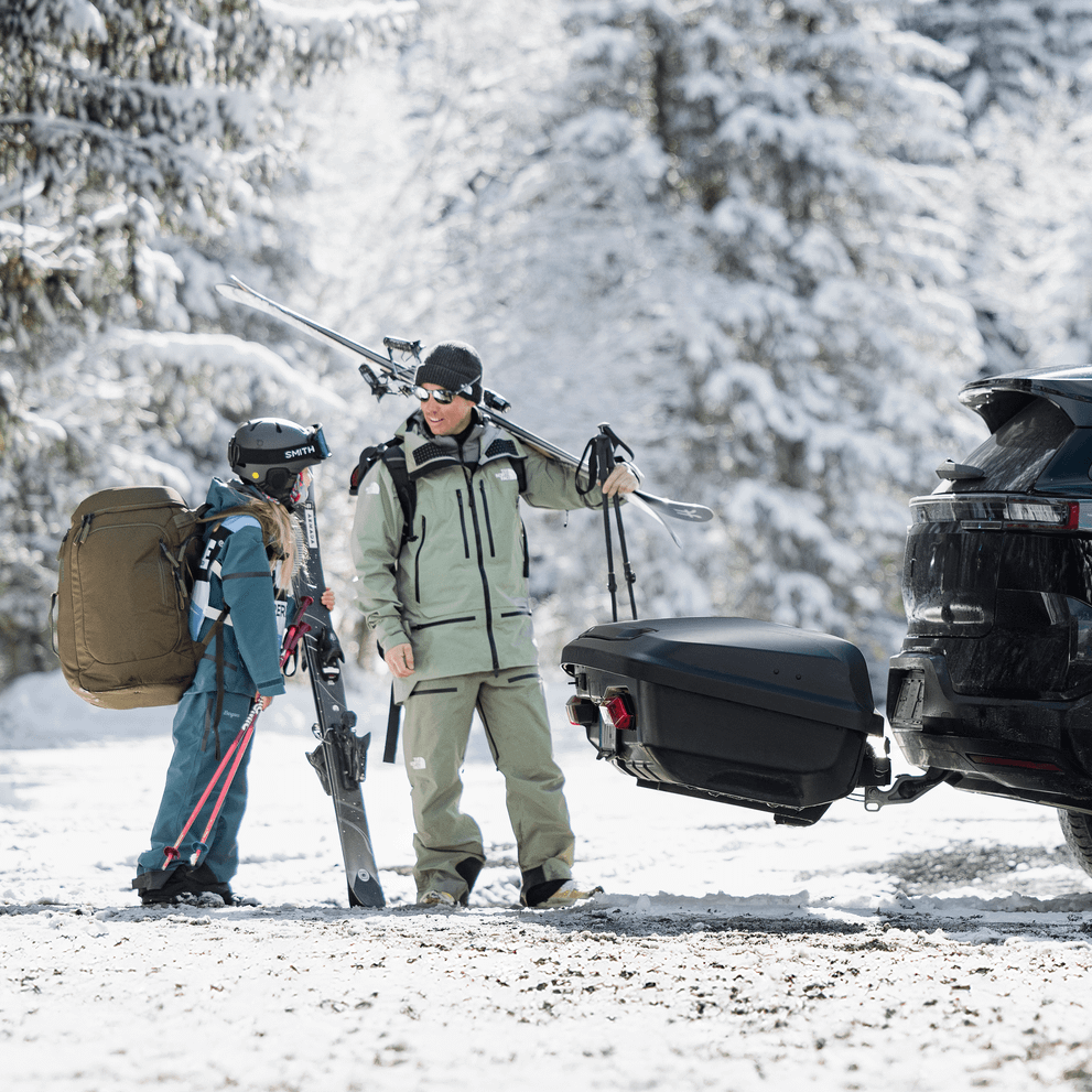 Two people dressed in ski gear with ski equipment, stand behind their car with a Thule Arcos XL mounted in a snowy forest setting.