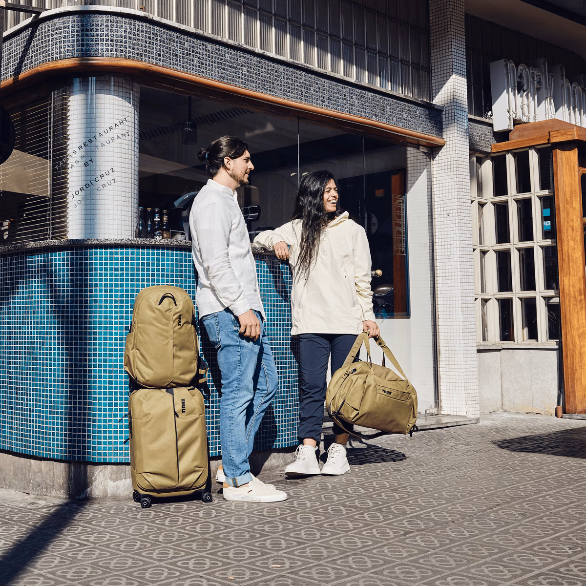A couple stand in the sun by a restaurant with tan bags including a Thule Aion Carry On Spinner.