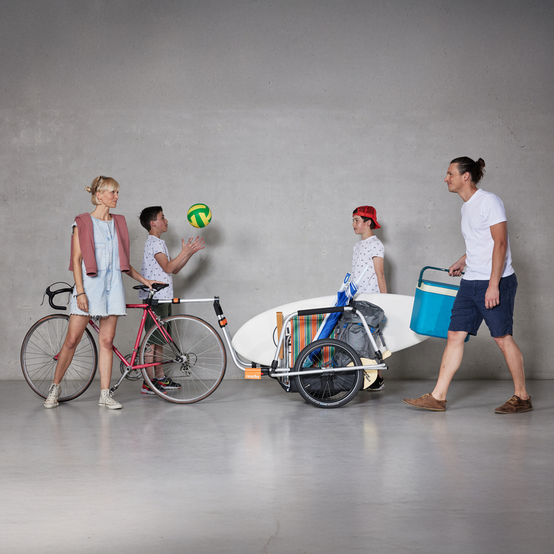 A family in a studio with a Reacha City bike trailer loaded with beach gear, including a volleyball and cooler, ready for an outing.