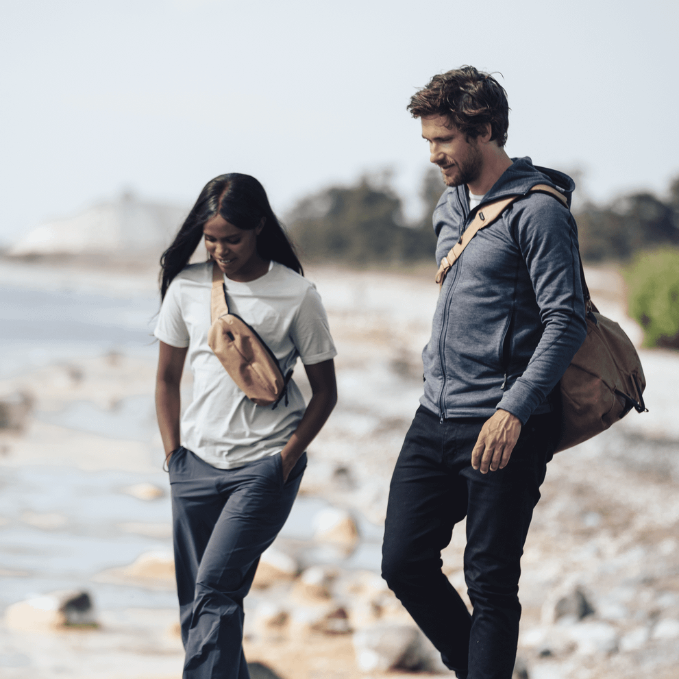 Two people walk down a beach, the man has a duffel and the woman has a Thule Aion sling bag.