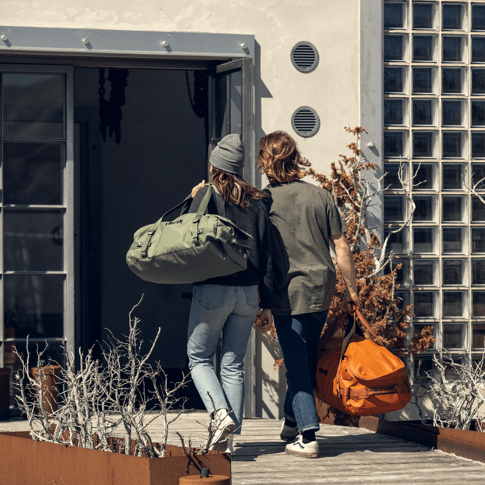 A couple walk into a building carrying a green and orange Thule Chasm duffel bags.