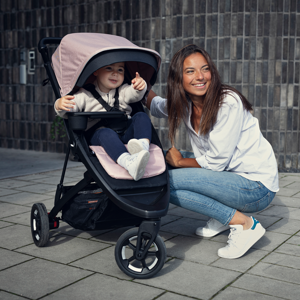 A woman in white kneels next to her baby in a pink stroller with a pink Thule Seat Liner.