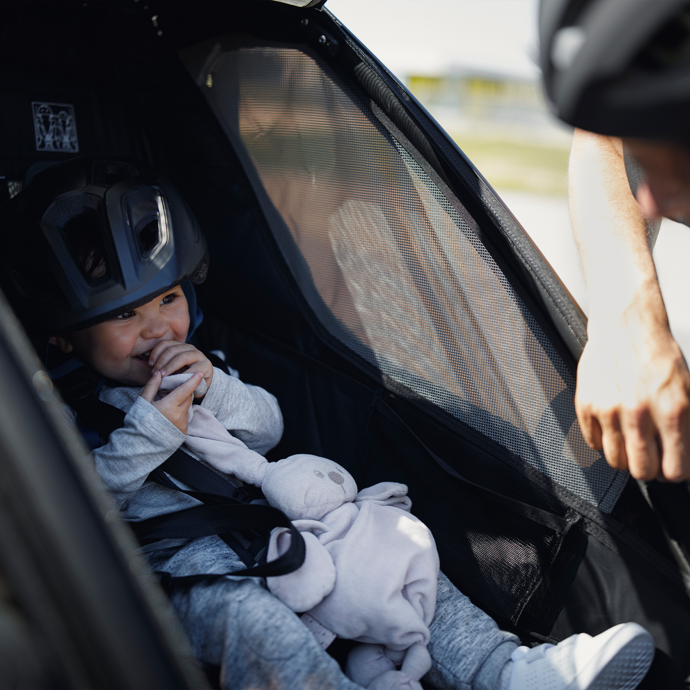 A close-up of a baby with a toy laughing inside a black Thule Chariot Sport kids' bike trailer.