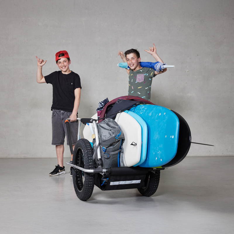 Two smiling young people in a studio showing a Reacha Sport Beach bike trailer loaded with a large blue paddleboard and camping gear.